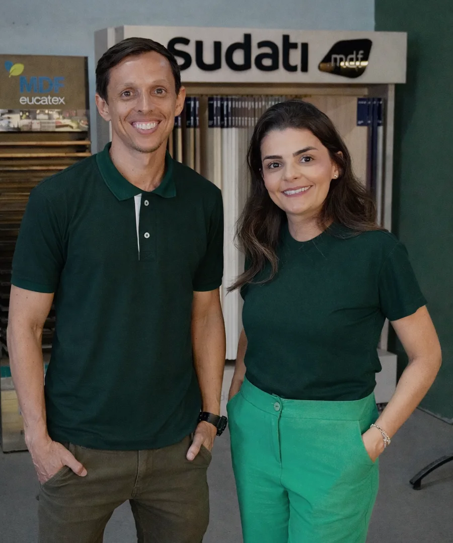 Otávio e Ana Paula, sócios da Santa Clara MDF, sorrindo na loja em frente ao expositor de chapas